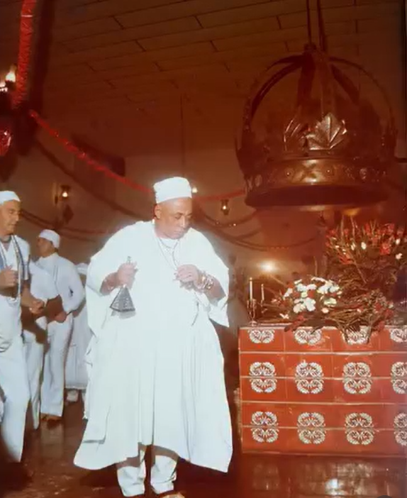 Pai Caio de Xangô em cerimônia: babalorixá de branco com agogô e fios de contas, ao lado de altar com flores e velas e coroa de Xangô